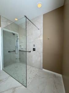 Spacious bathroom with a frameless glass shower enclosure featuring a minimalist design. The shower area has light marble-look tiles and a sleek, modern grab bar on the wall, complemented by a soft beige accent wall.