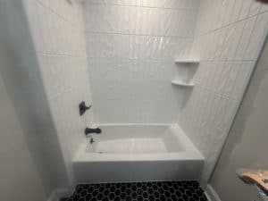 Bathroom with a white tub and shower area featuring textured white wall tiles, built-in corner shelves, and a contrasting black hexagonal tile floor, creating a modern and stylish design.