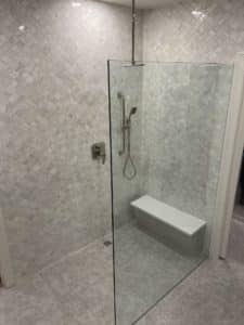 Elegant shower with a frameless glass enclosure and a built-in floating bench. The walls and floor are covered in light gray mosaic tiles, creating a cohesive, textured look. A sleek handheld showerhead completes the modern design.