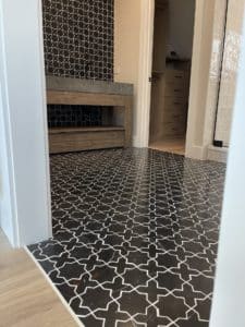 Bathroom with bold black-and-white patterned floor tiles featuring an intricate geometric design. The vanity area has a natural wood finish, adding warmth to the space. A dark tiled accent wall complements the floor for a cohesive look.