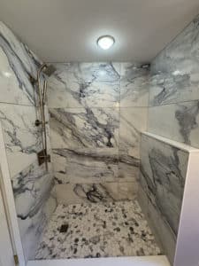 Luxurious shower with large, dramatic marble-look tiles on the walls, featuring bold gray veining. The shower floor has a pebble stone design, adding texture and a natural feel to the space.